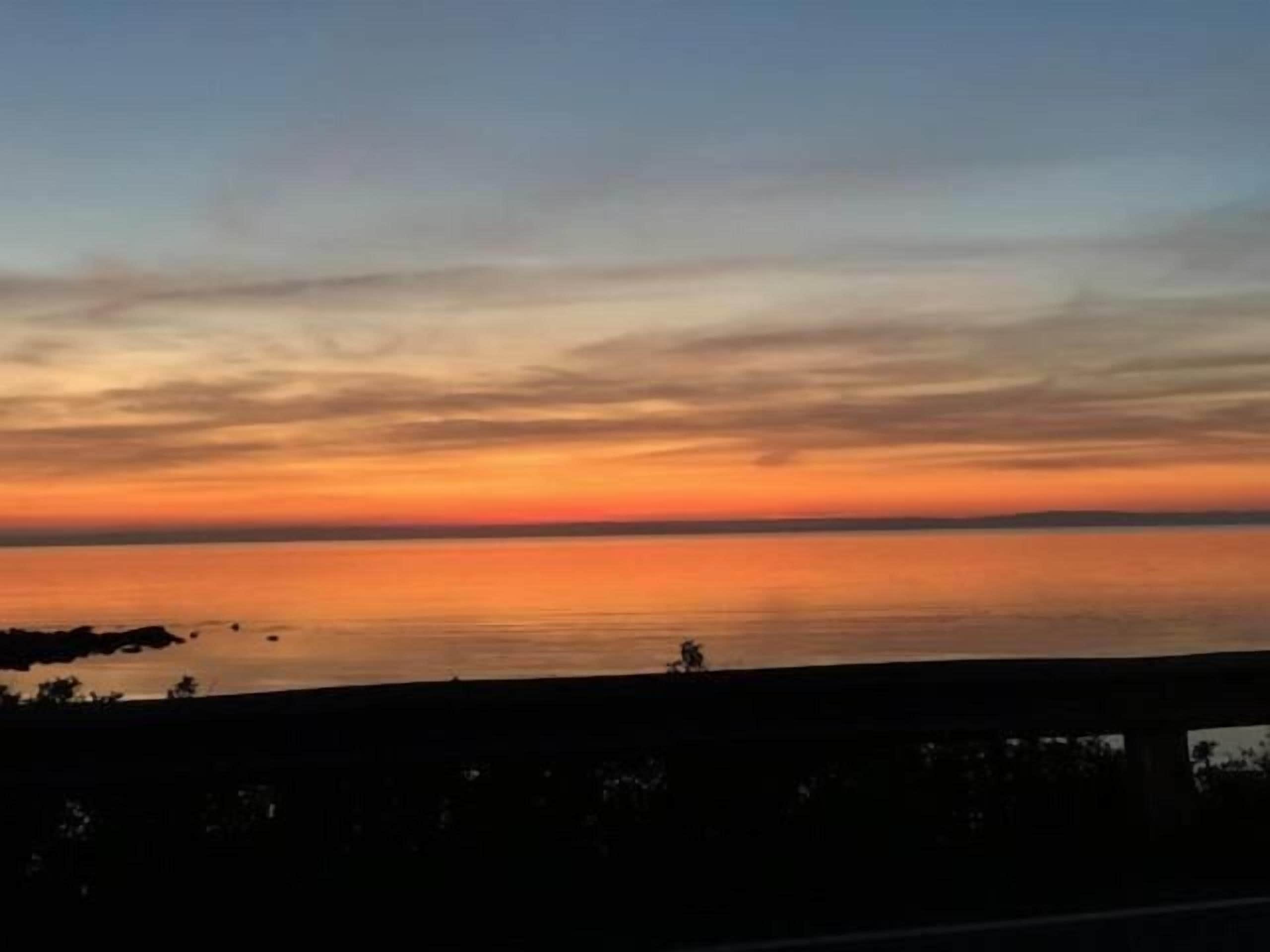A sunset over a calm body of water with a silhouette of a bridge and trees in the foreground.