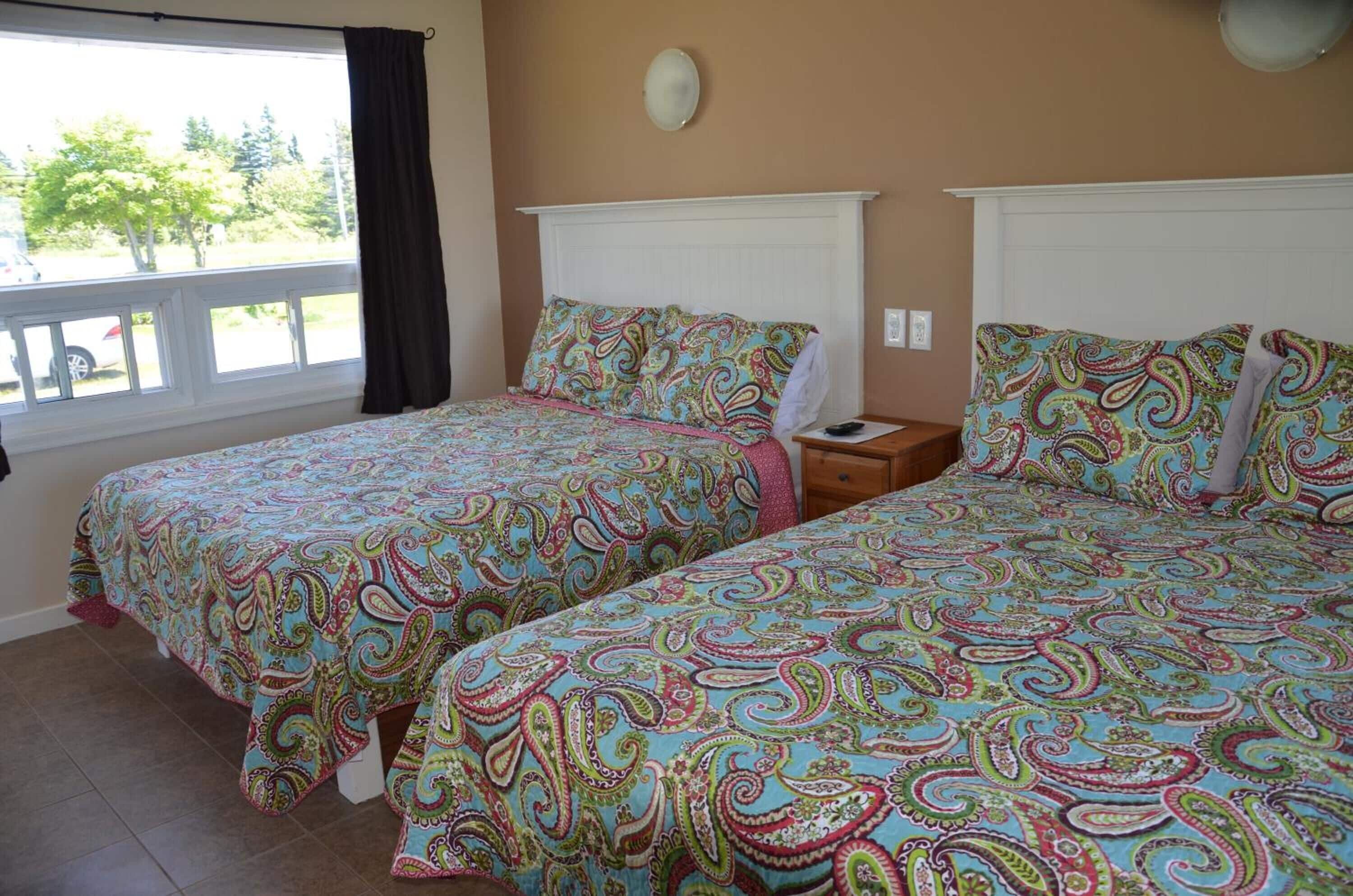 Habitación con dos camas, cada una con una colorida colcha de estampado paisley, una mesita de noche de madera con lámpara, y una ventana con vistas a árboles y un coche.