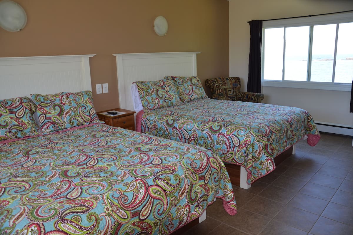 A room with two beds, each with a patterned bedspread, a nightstand, and a window with a view of the sea.