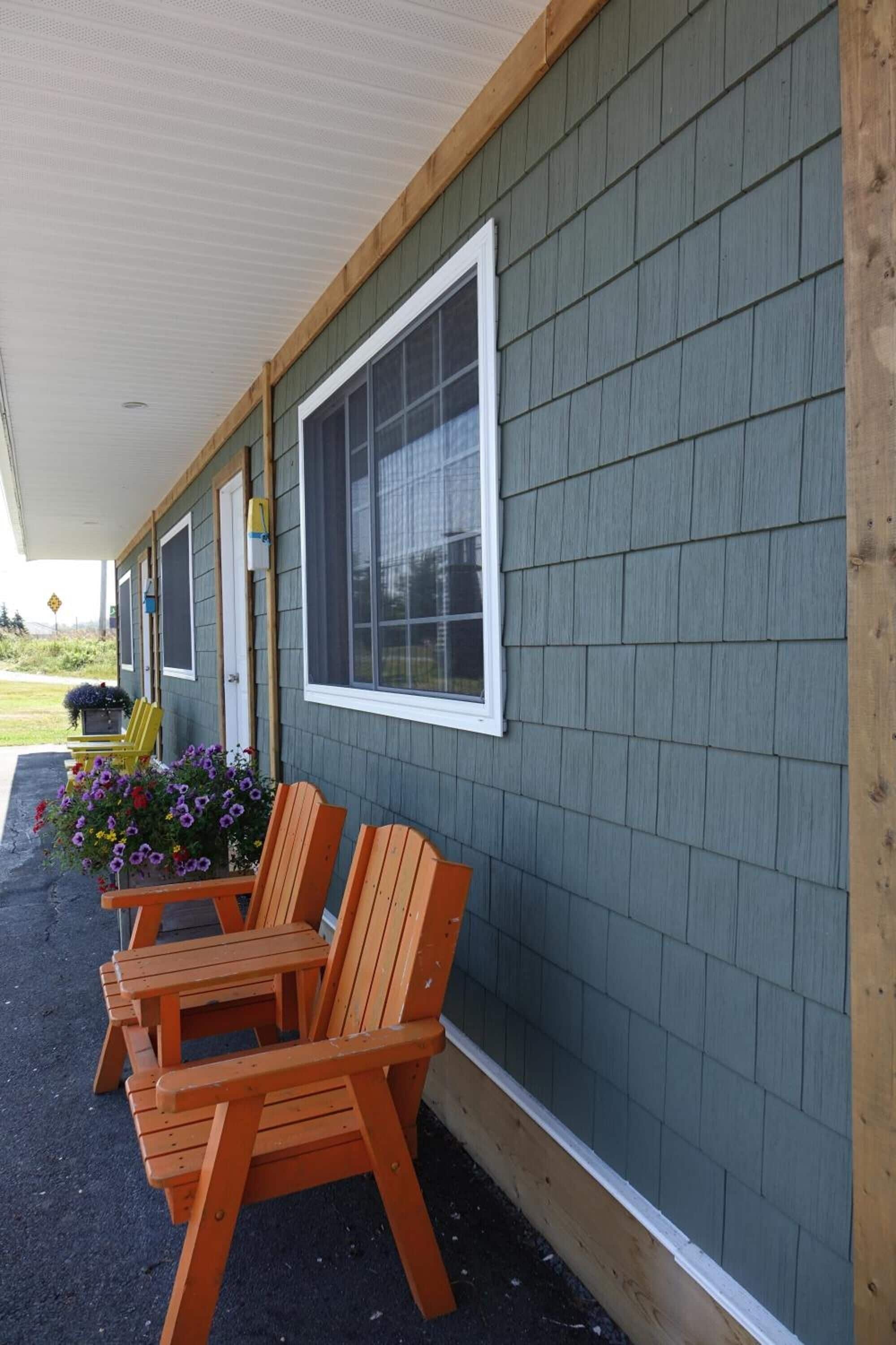A row of wooden chairs outside a building with a green siding exterior and a white window.