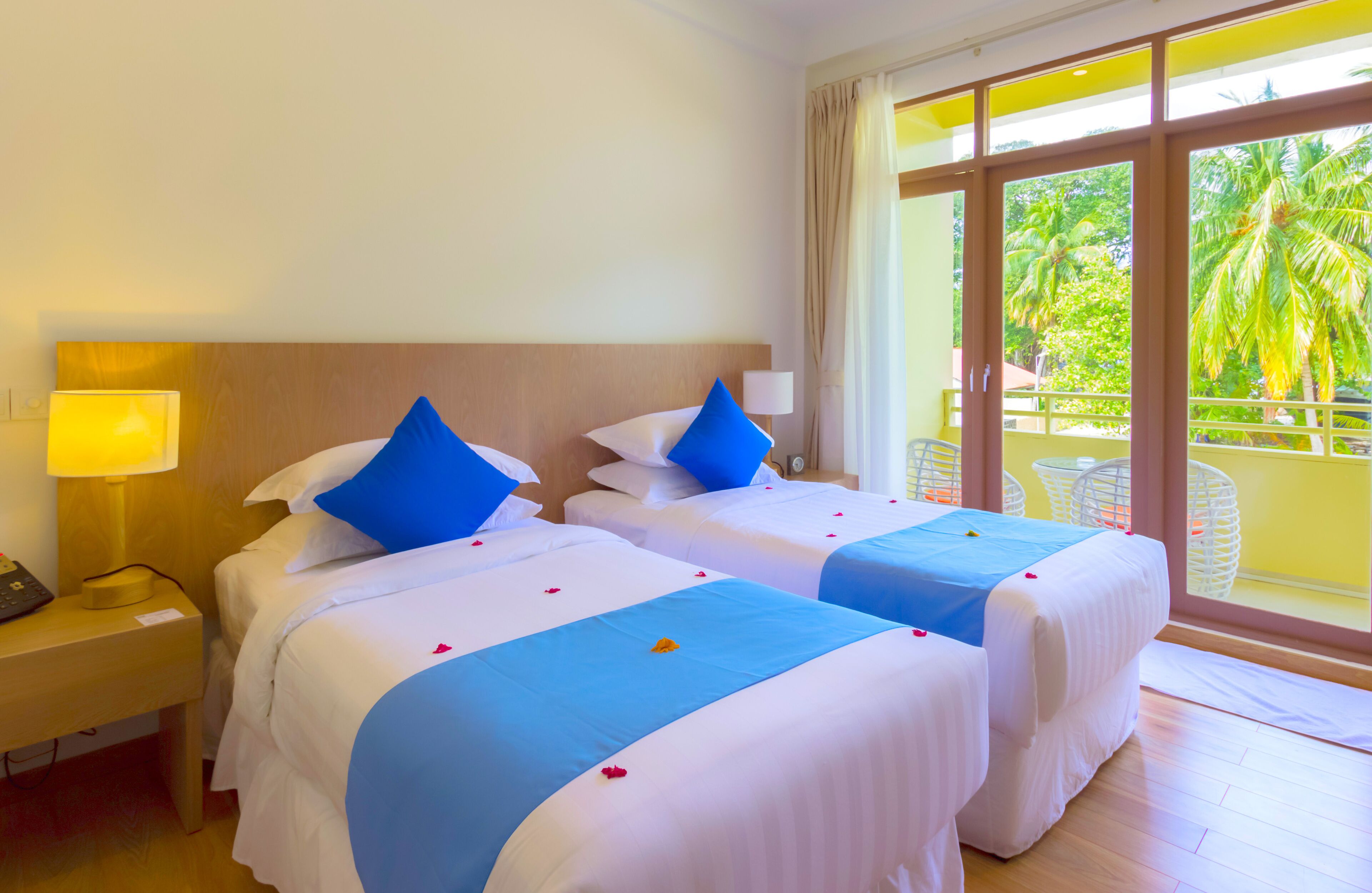 A hotel room with two beds, a nightstand with a lamp, and a balcony with a view of palm trees.