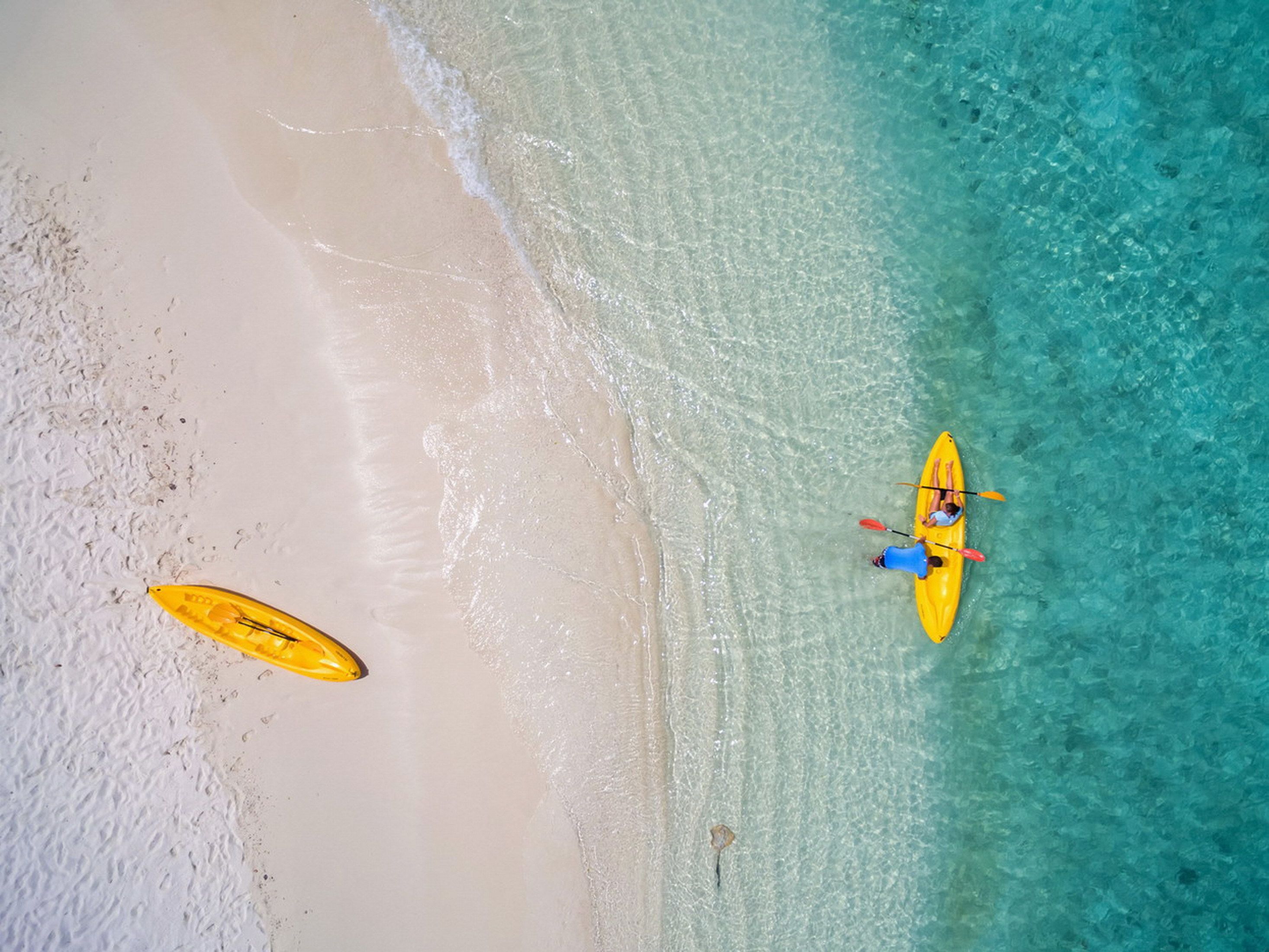 An aerial view of a beach with clear turquoise water, a sandy shore, and a yellow kayak with two people paddling.