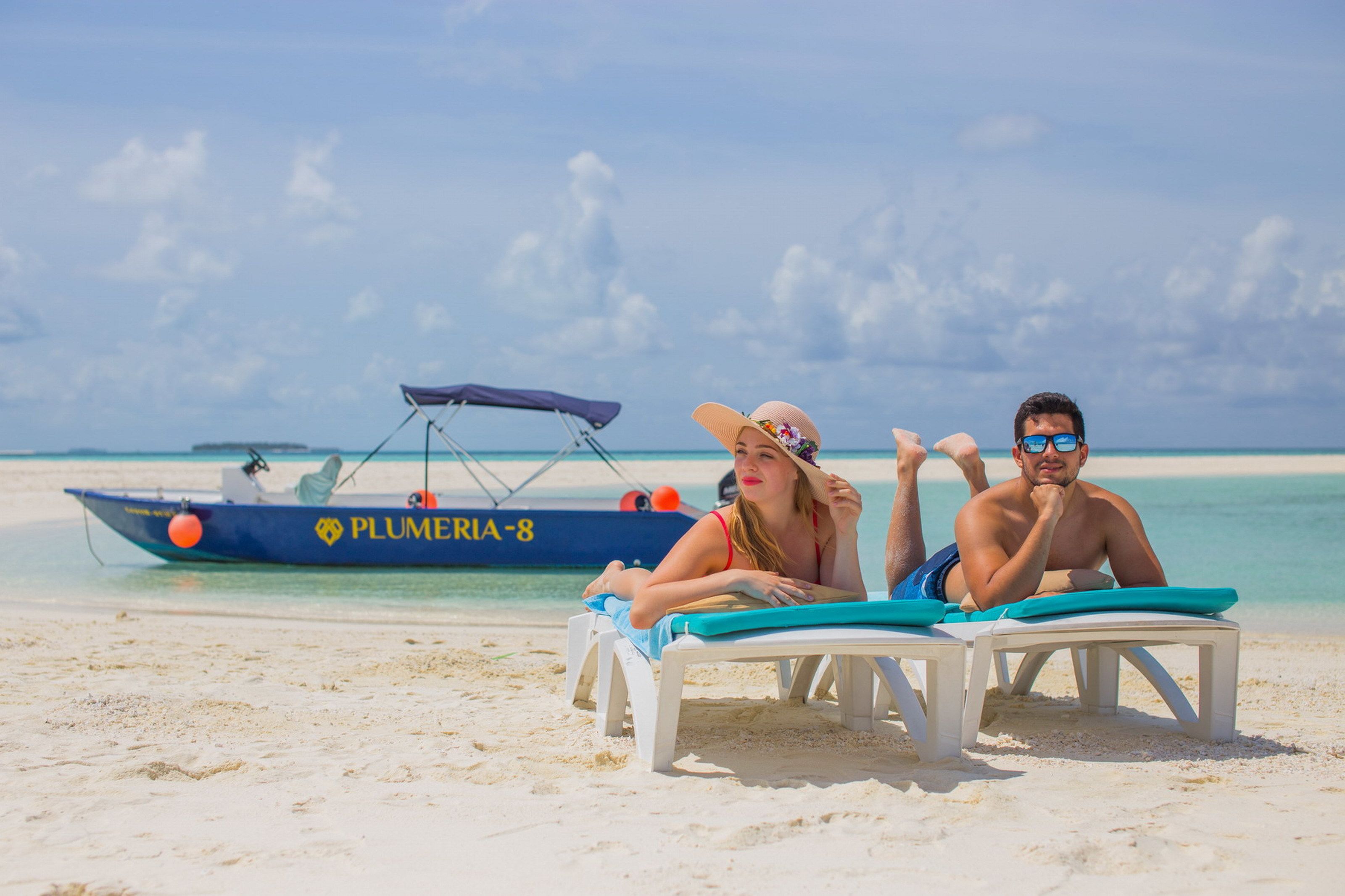 Two people relaxing on beach chairs with a boat named 'PLUMERIA-8' in the background.