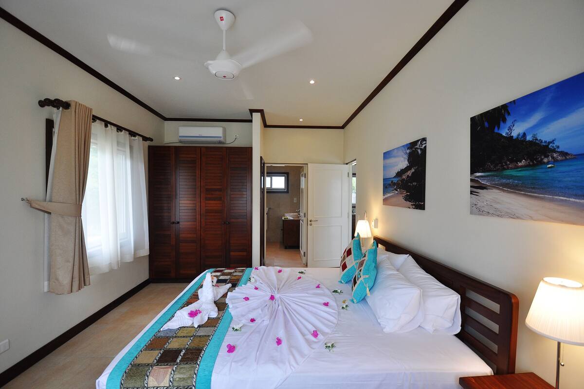 A hotel room with a bed, a ceiling fan, a painting of a beach, and a door leading to a bathroom.