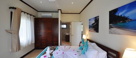 A hotel room with a bed, a ceiling fan, a painting of a beach, and a door leading to a bathroom.