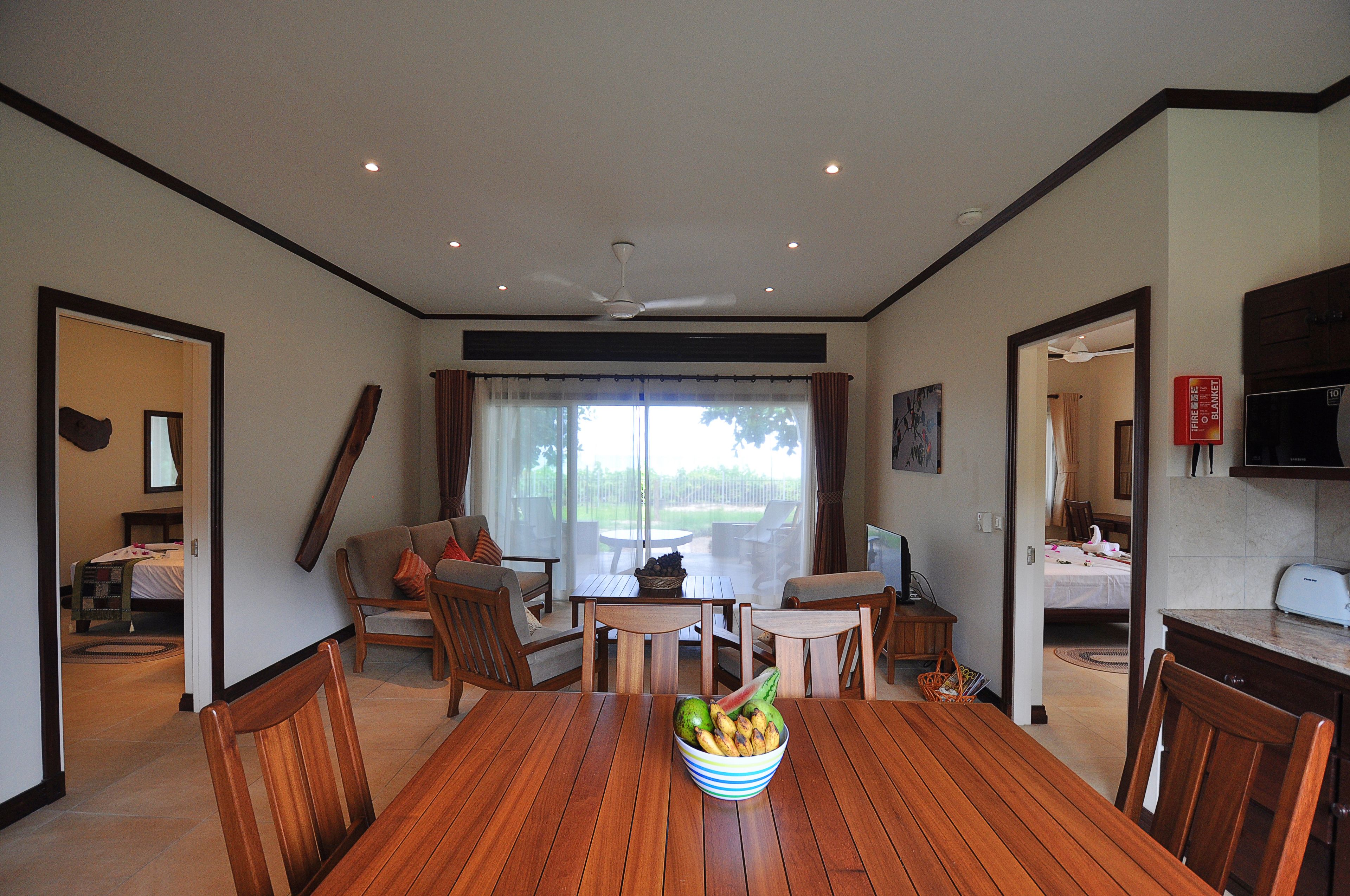 A dining area with a wooden table, chairs, and a bowl of fruit. There is a ceiling fan, a window with curtains, and a kitchen area visible in the background.