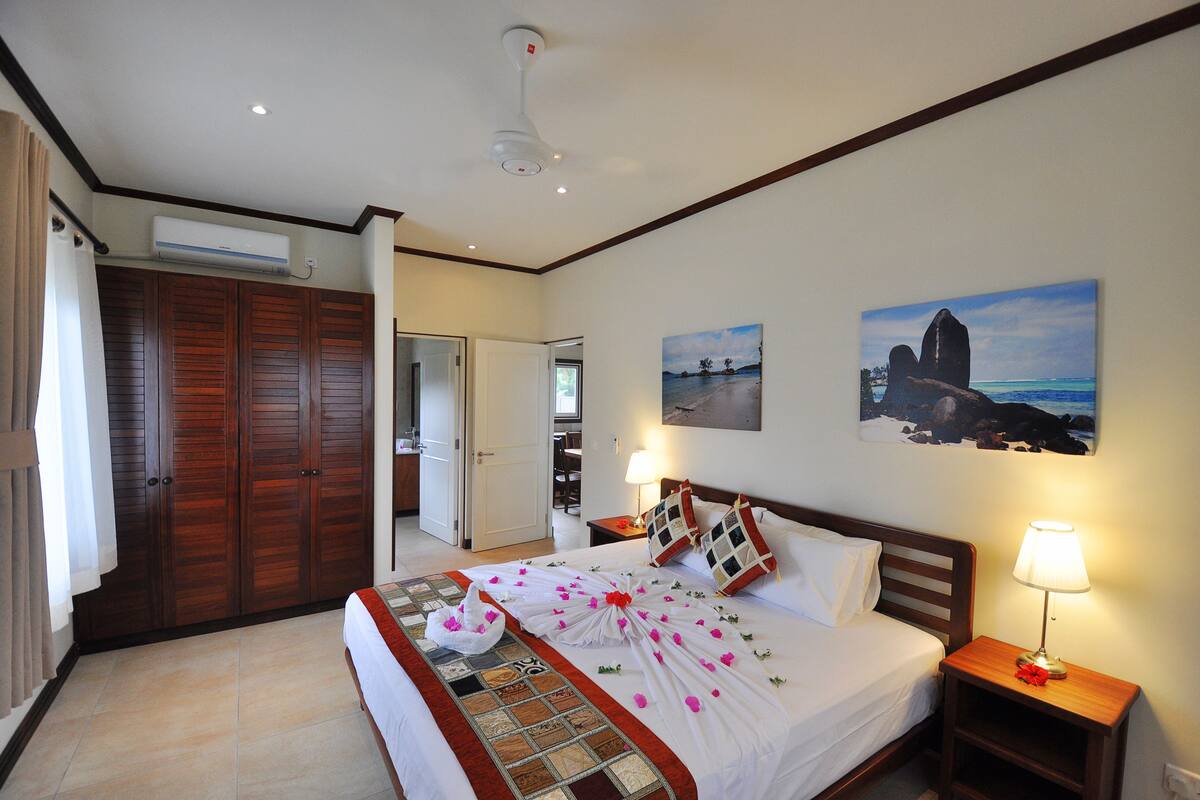 A hotel room with a bed, a nightstand, a wall-mounted air conditioner, and a door leading to another room.