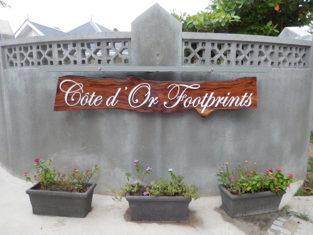 Una señal con el texto 'Cote d'Or Footprints' sobre una pared gris, flanqueada por tres macetas con flores.