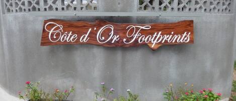 Una señal con el texto 'Cote d'Or Footprints' sobre una pared gris, flanqueada por tres macetas con flores.