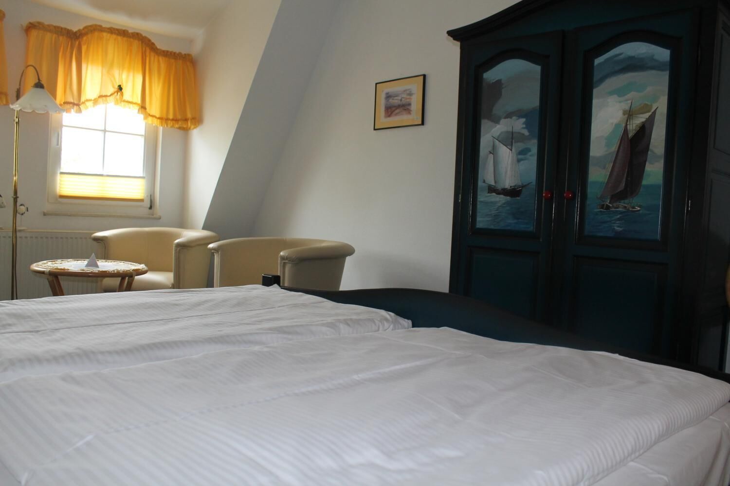A hotel room with a large bed, two armchairs, a small table, and a dark wooden wardrobe with a sailboat painting.