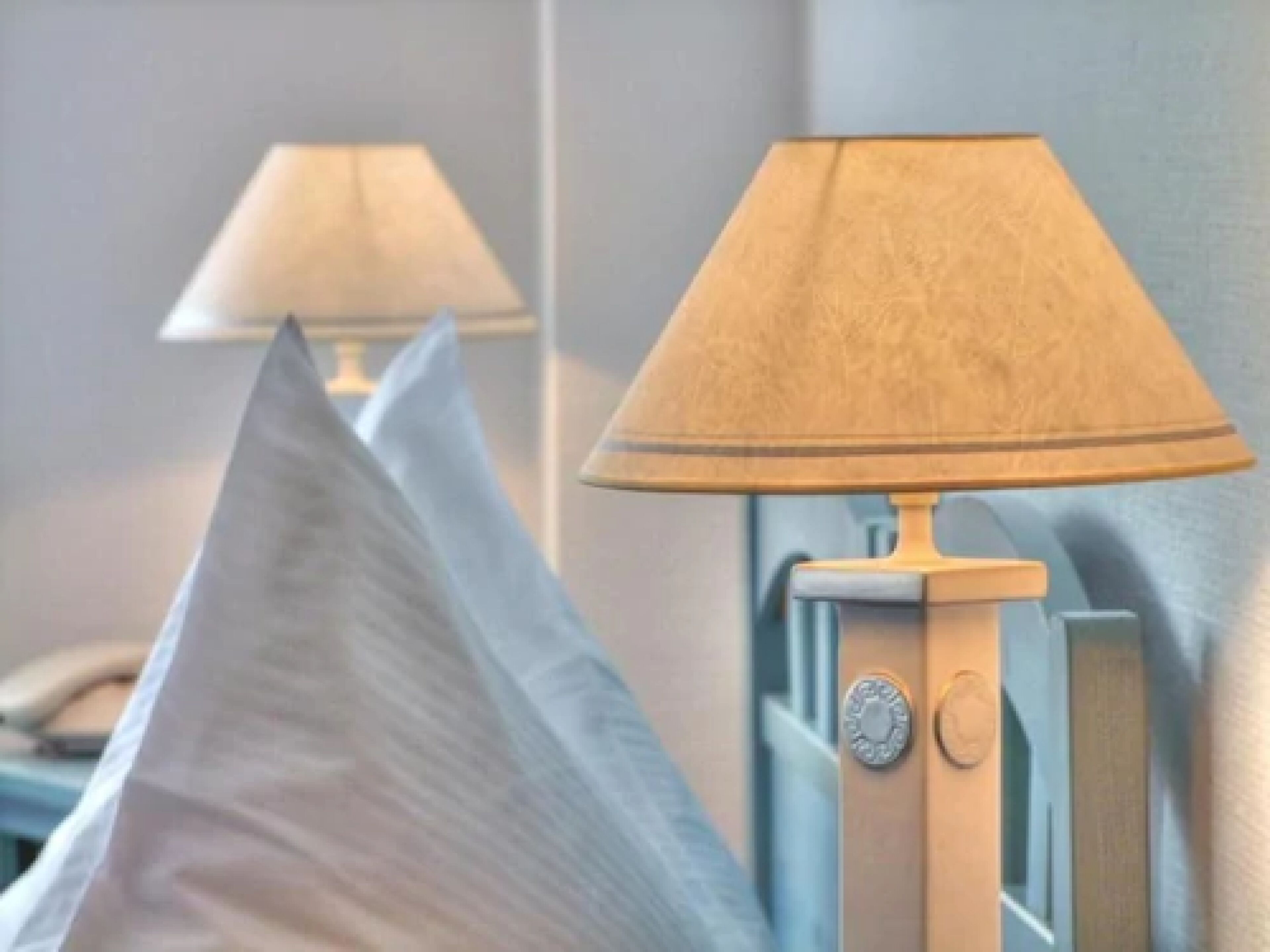 Une lampe dotée d’un abat-jour beige et d’un pied métallique, placée à côté d’un lit sur lequel se trouve un oreiller blanc.