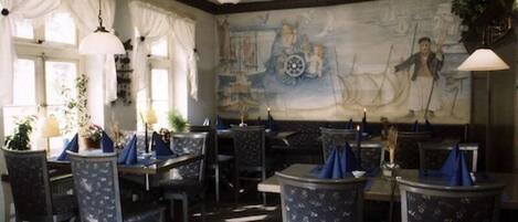 A restaurant with a nautical-themed mural, multiple tables with blue tablecloths, and hanging pendant lights.