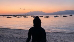 Eine Statue einer sitzenden Person am Strand, mit Booten im Wasser und Bergen im Hintergrund.