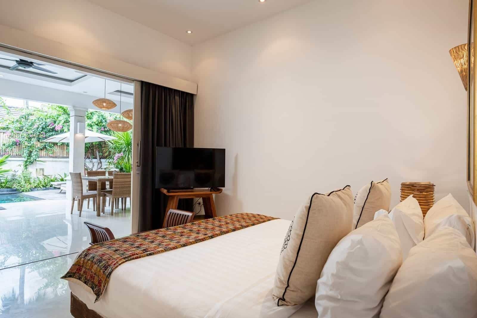 Una camera d'albergo moderna con un letto grande, una TV a schermo piatto su un supporto in legno, un'area pranzo con mobili in vimini e vista su un giardino attraverso una porta in vetro.