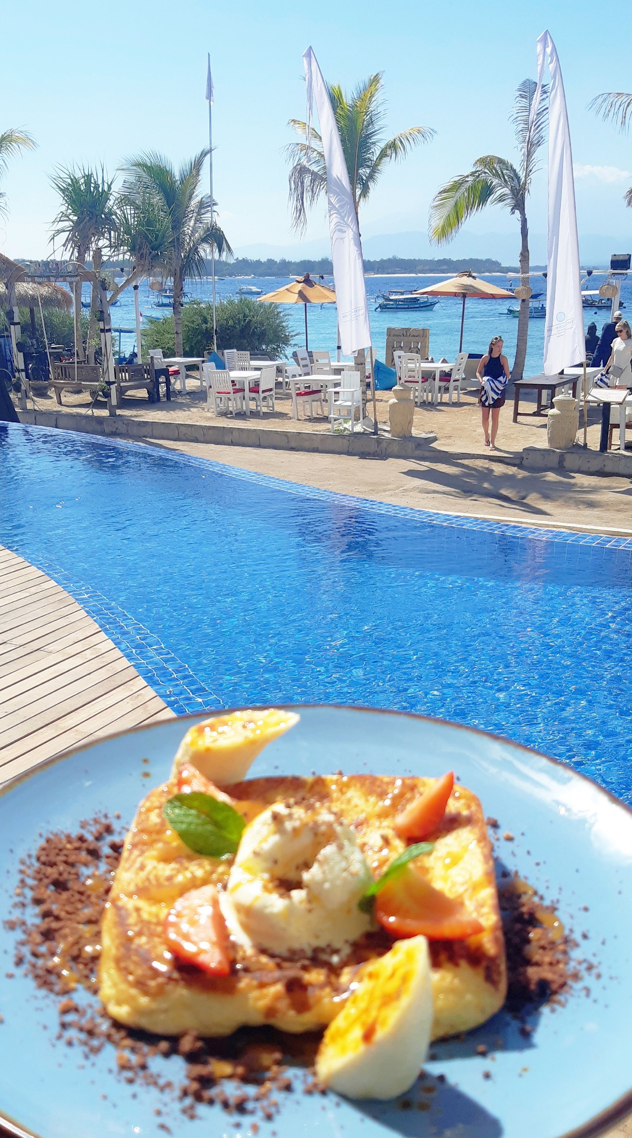 A poolside dining area with a view of the sea and boats, a table with a plate of food, and a wooden deck.