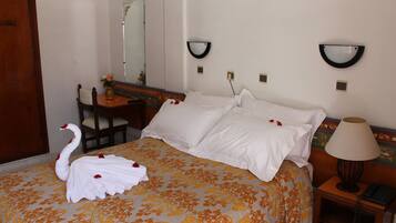 Una habitación de hotel con una cama, una silla de madera, un escritorio, un espejo y una mesita de noche con una lámpara.