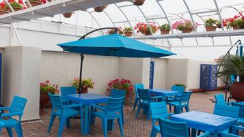 An outdoor patio area with blue tables and chairs, surrounded by potted plants and a blue umbrella.