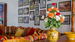 A room with a colorful patterned sofa, a wooden table with a yellow vase and a bowl, and a wall decorated with framed posters. - Hotel Sindibad (Agadir)