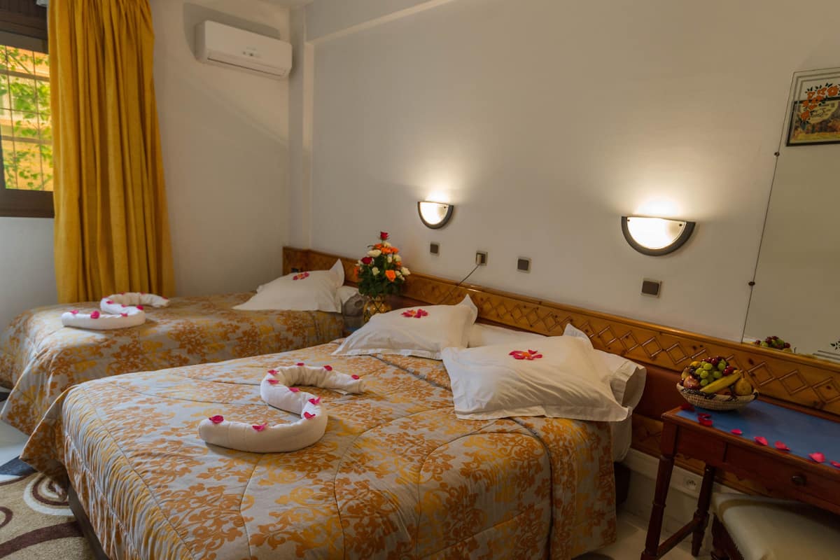 Habitación de hotel con dos camas, cada una con ropa de cama y toallas con motivos florales. Hay lámparas fijadas a la pared y una mesita con un frutero.