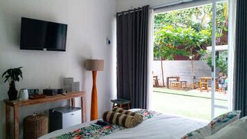 A hotel room with a bed, a desk with a flat-screen TV, a small refrigerator, a potted plant, and a view of an outdoor patio with a table and chairs.