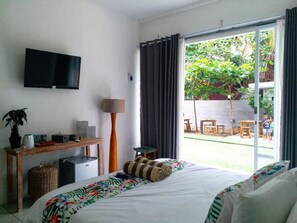 A hotel room with a bed, a desk with a flat-screen TV, a small refrigerator, a potted plant, and a view of an outdoor patio with a table and chairs.
