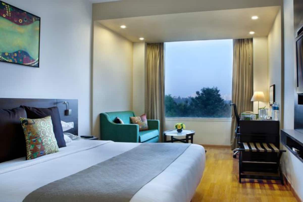 A hotel room with a bed, a green armchair, a small table, a TV, and a window with curtains.