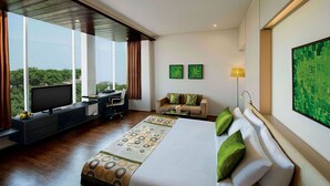 A modern hotel room with a large bed, a desk, and a view of greenery through large windows. - Lemon Tree Hotel Vadodra (Vadodara)