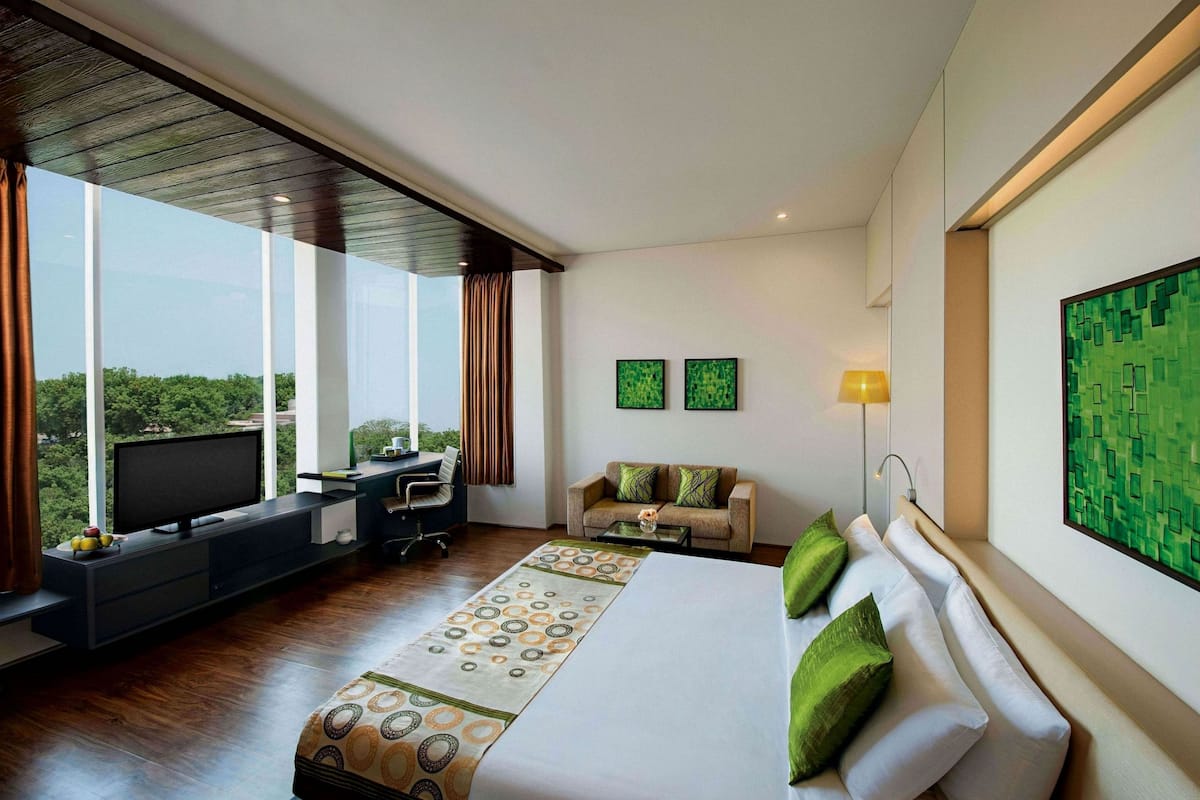 A modern hotel room with a large bed, a desk, and a view of greenery through large windows.