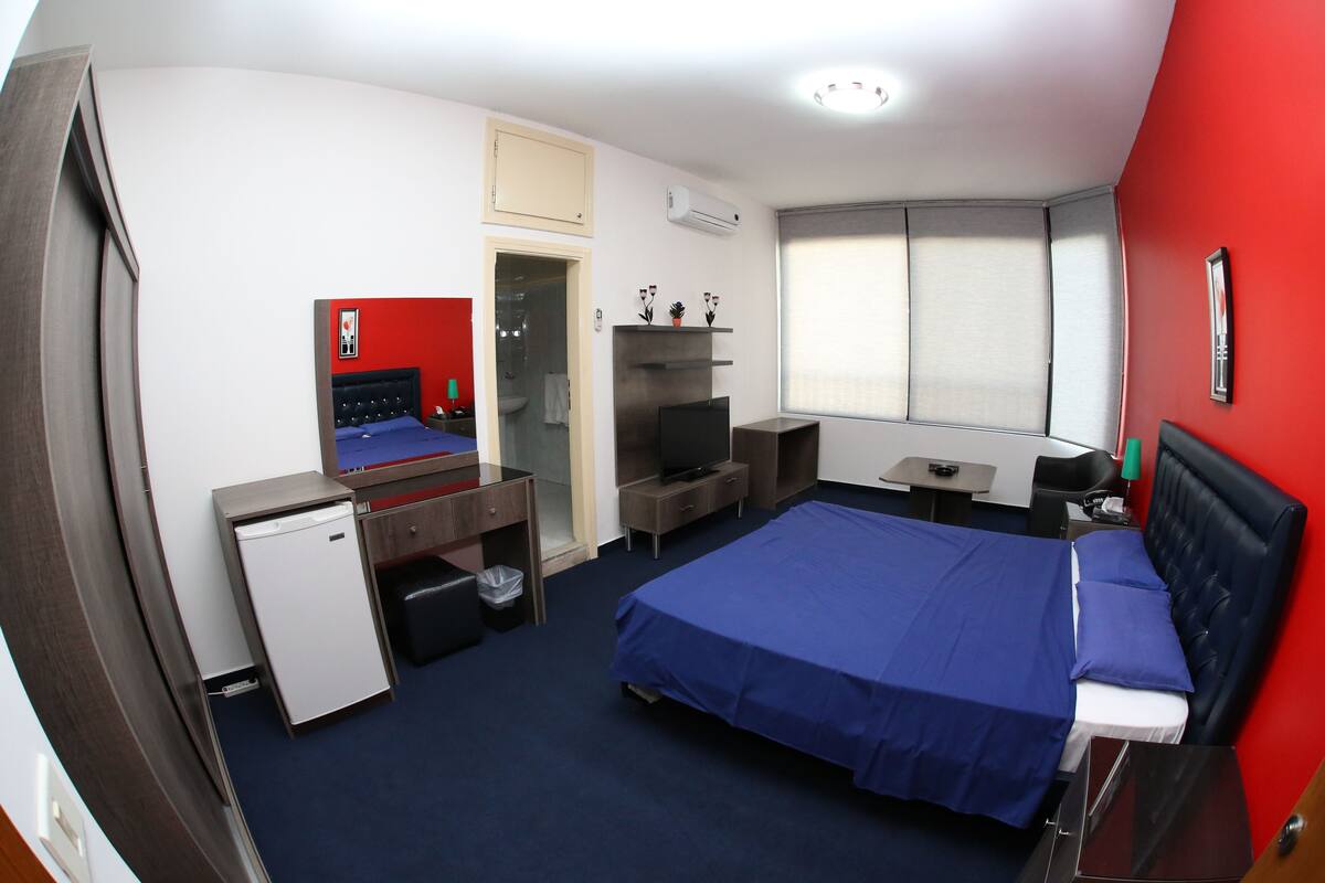 A hotel room with a bed, a desk, a small fridge, a TV, and a window with blinds.