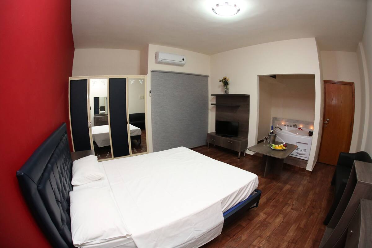 A hotel room with a bed, a TV, a desk with a chair, a mirror, and a door leading to a bathroom.