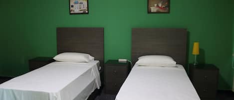 Dos camas individuales con sábanas blancas, una pared verde y una mesita de noche con lámpara.