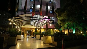 Entrada de hotel con paseo techado, varias banderas y plantas en macetas.