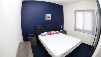 A bedroom with a bed, a nightstand, a bedside table, a wall-mounted lamp, and a window with blinds.