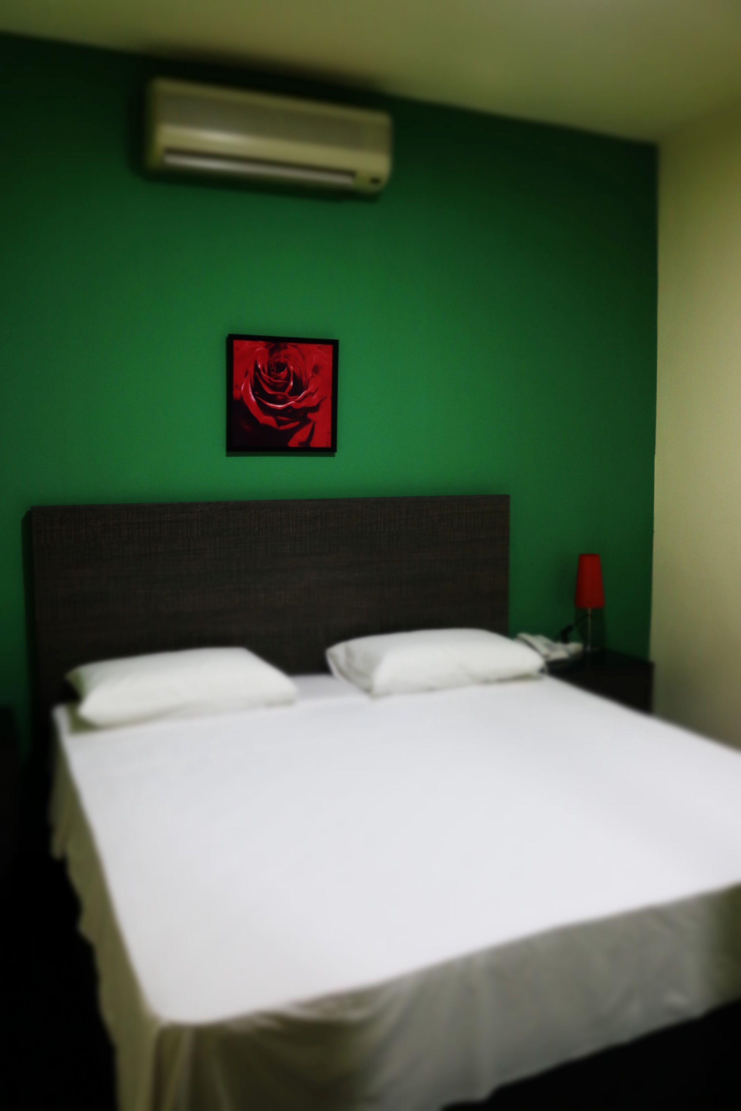 A bed with white linens, a dark headboard, and a red rose artwork on the green wall.