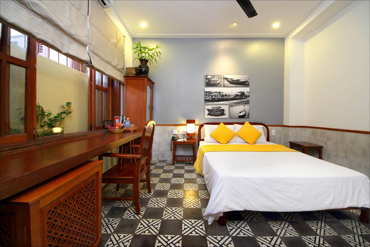 A hotel room with a bed, a desk, a chair, a nightstand, a lamp, and a wall with framed pictures.