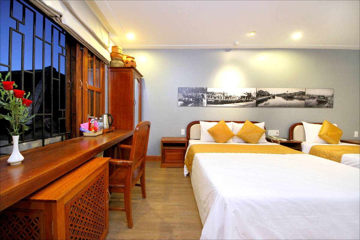 A hotel room with a wooden desk, a chair, two beds with white and yellow bedding, a nightstand, and a wall with three black and white photographs.