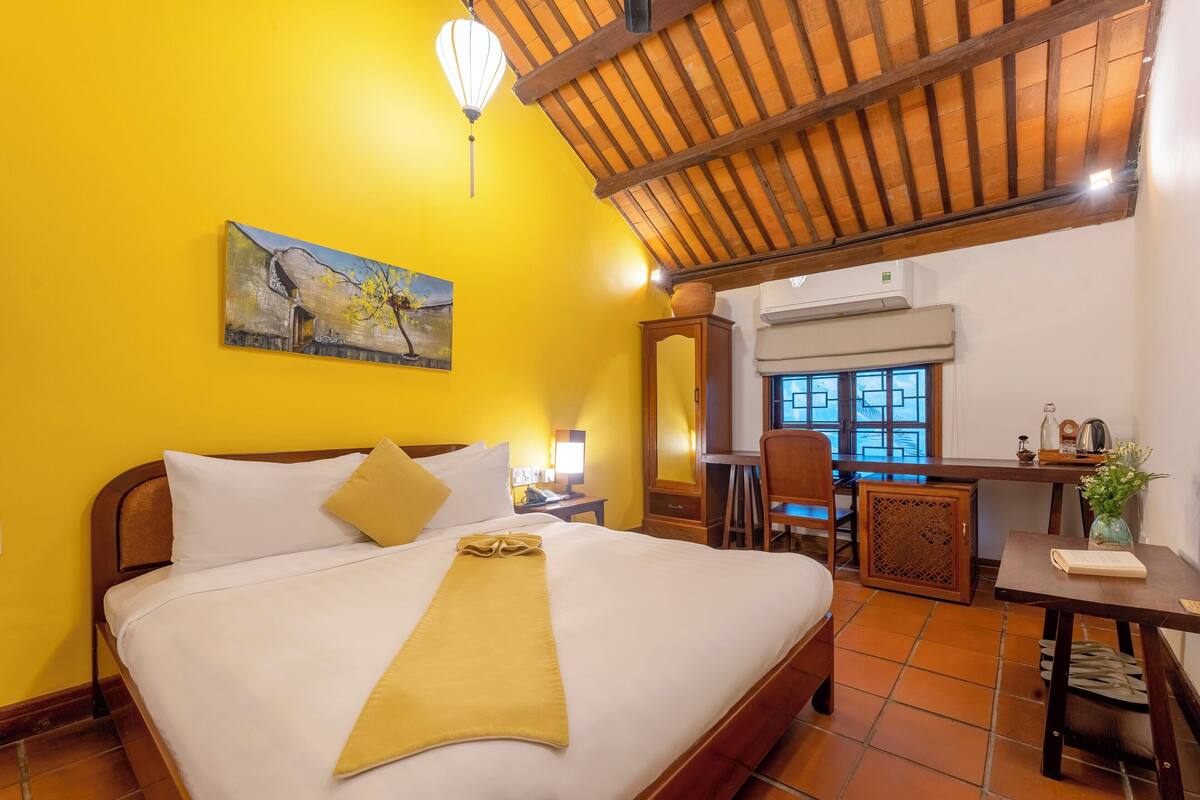 A hotel room with a bed, a desk, a chair, a window, and a painting on the wall.