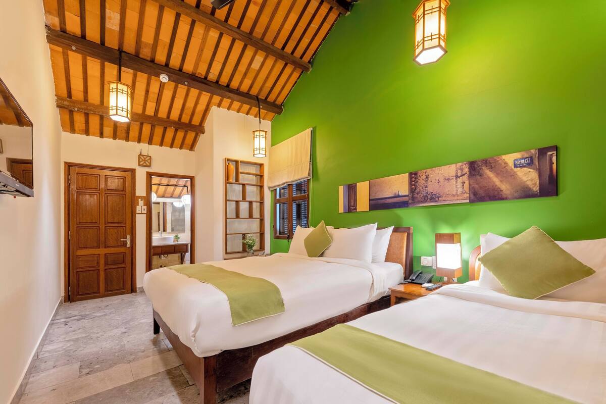 A hotel room with two beds, wooden beams on the ceiling, a green wall, and a painting above the beds.