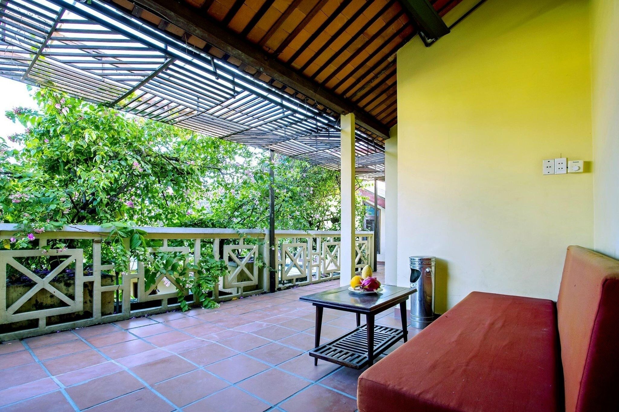 A covered outdoor area with a red sofa, a table with fruits, and a view of greenery.
