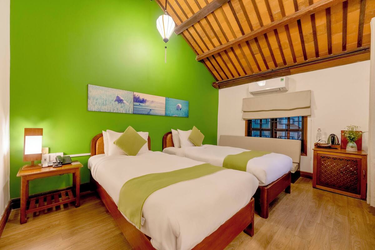 A hotel room with two beds, a wooden nightstand, a lamp, and a painting on the wall.
