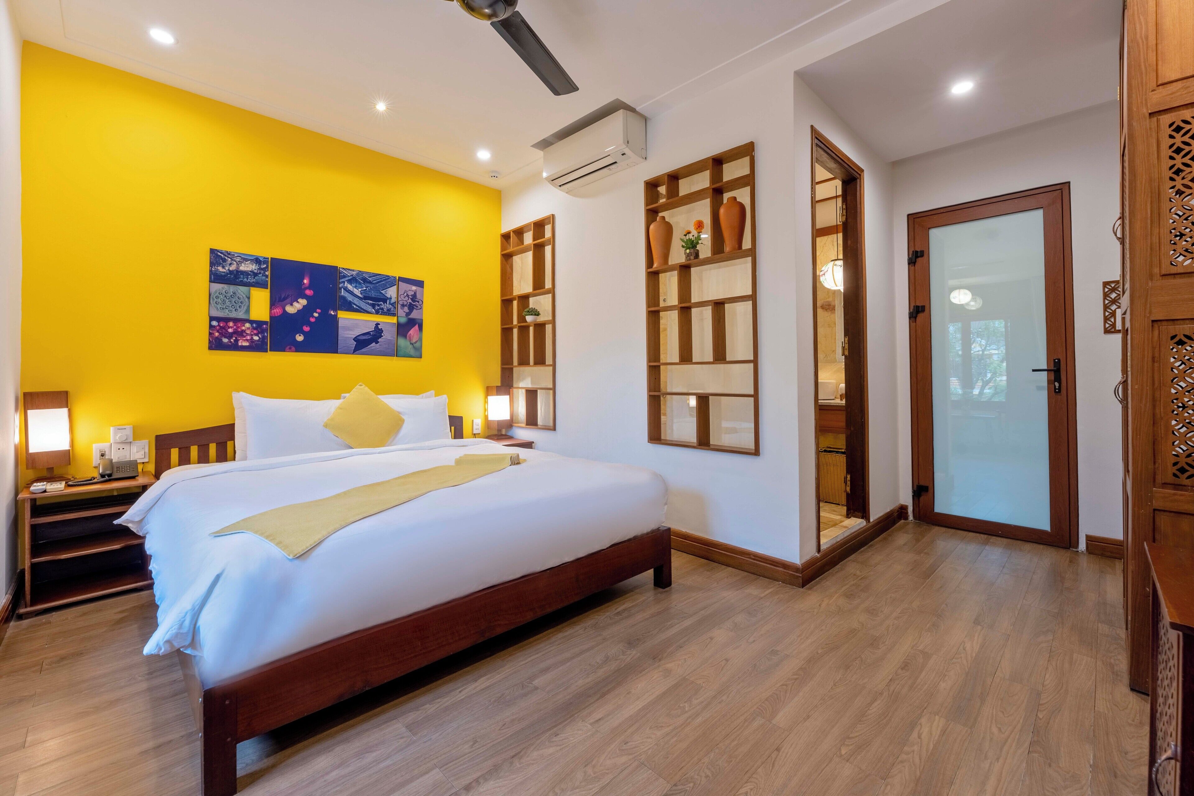 A hotel room with a large bed, a yellow accent wall, a wooden floor, a built-in shelf with decorative items, and a sliding glass door leading to a balcony.