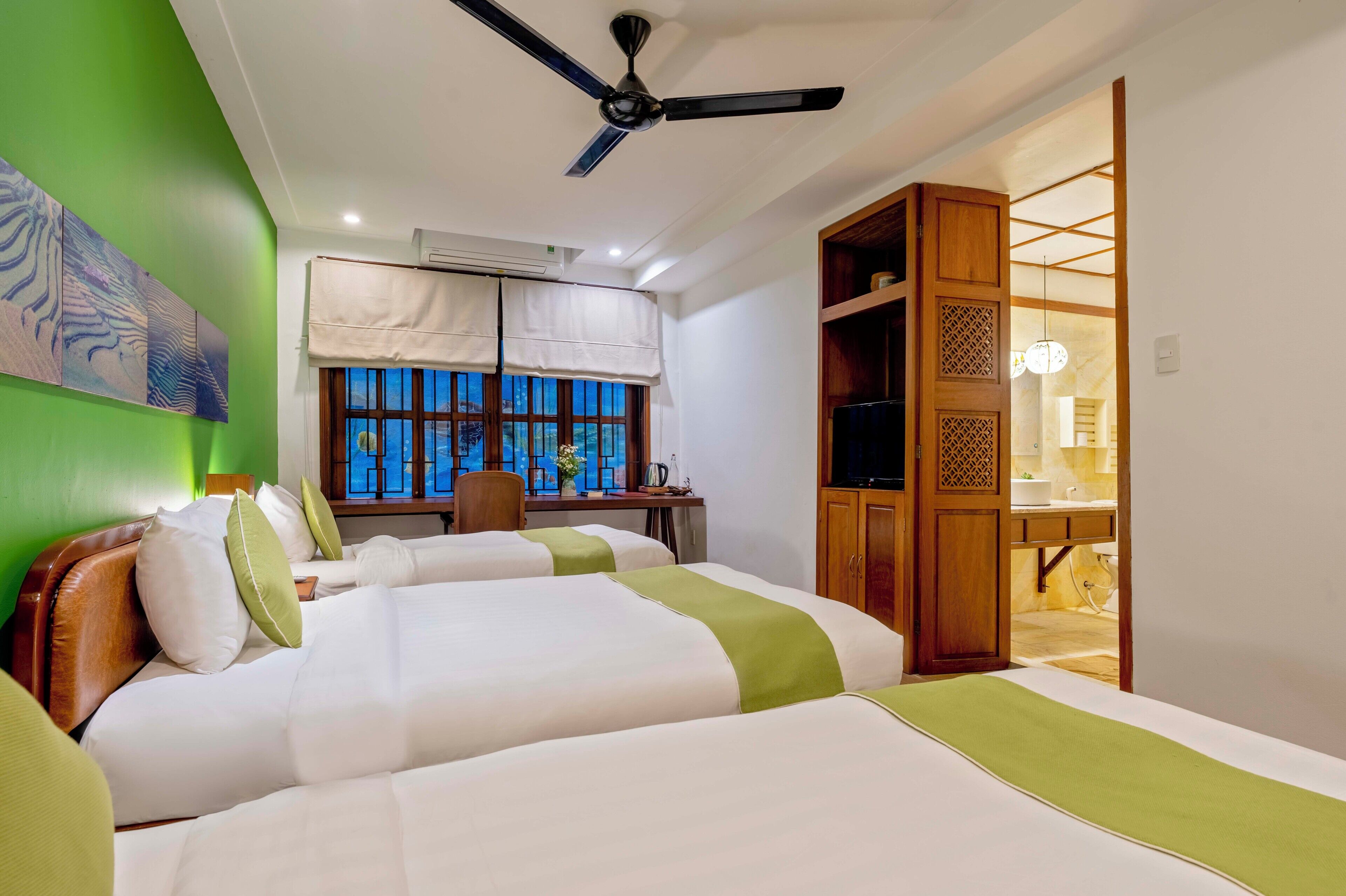 A hotel room with two beds, a desk, a chair, a ceiling fan, and a view of the outside through a window.