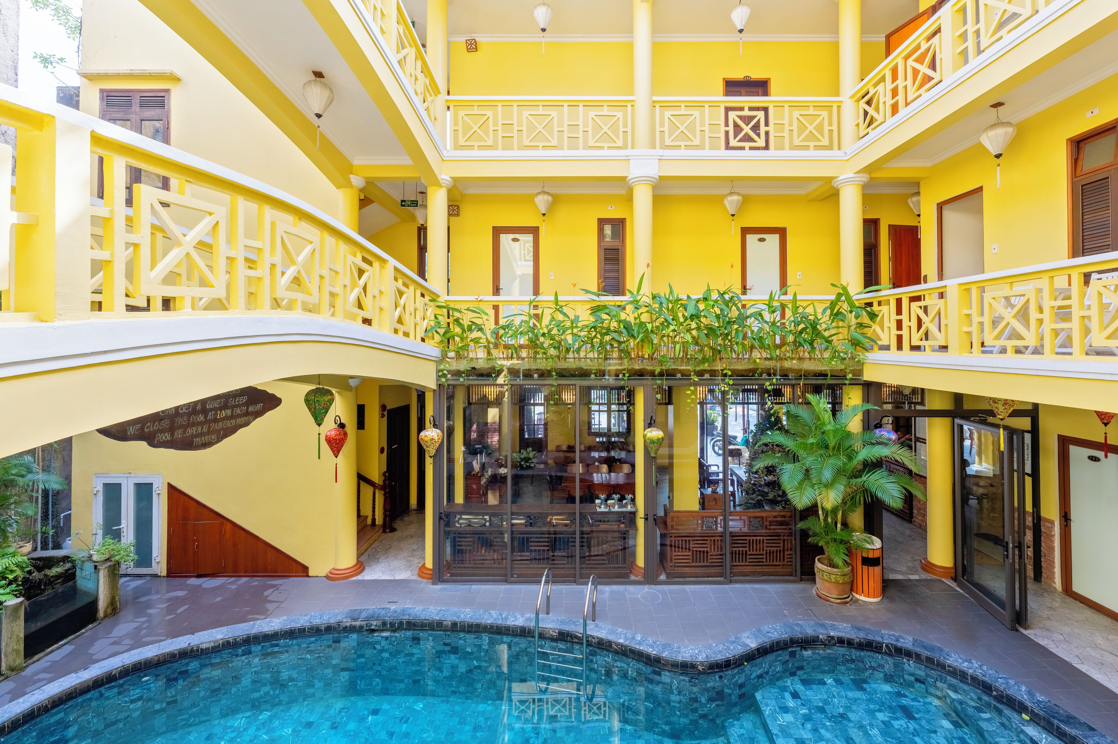 A multi-level indoor swimming pool with yellow walls and white railings, surrounded by potted plants and furniture.