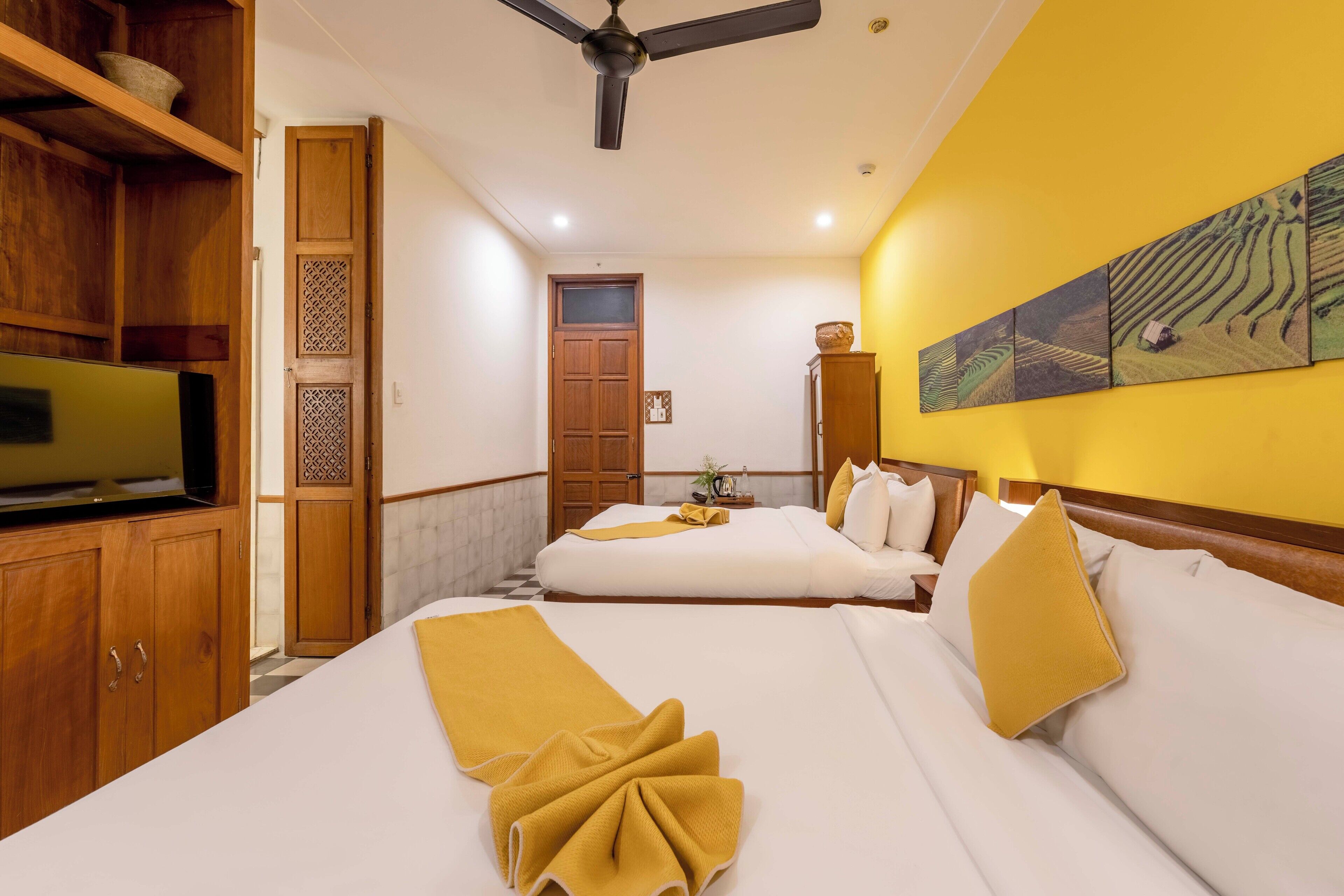 A hotel room with two beds, wooden furniture, a ceiling fan, and a wall with a landscape painting.