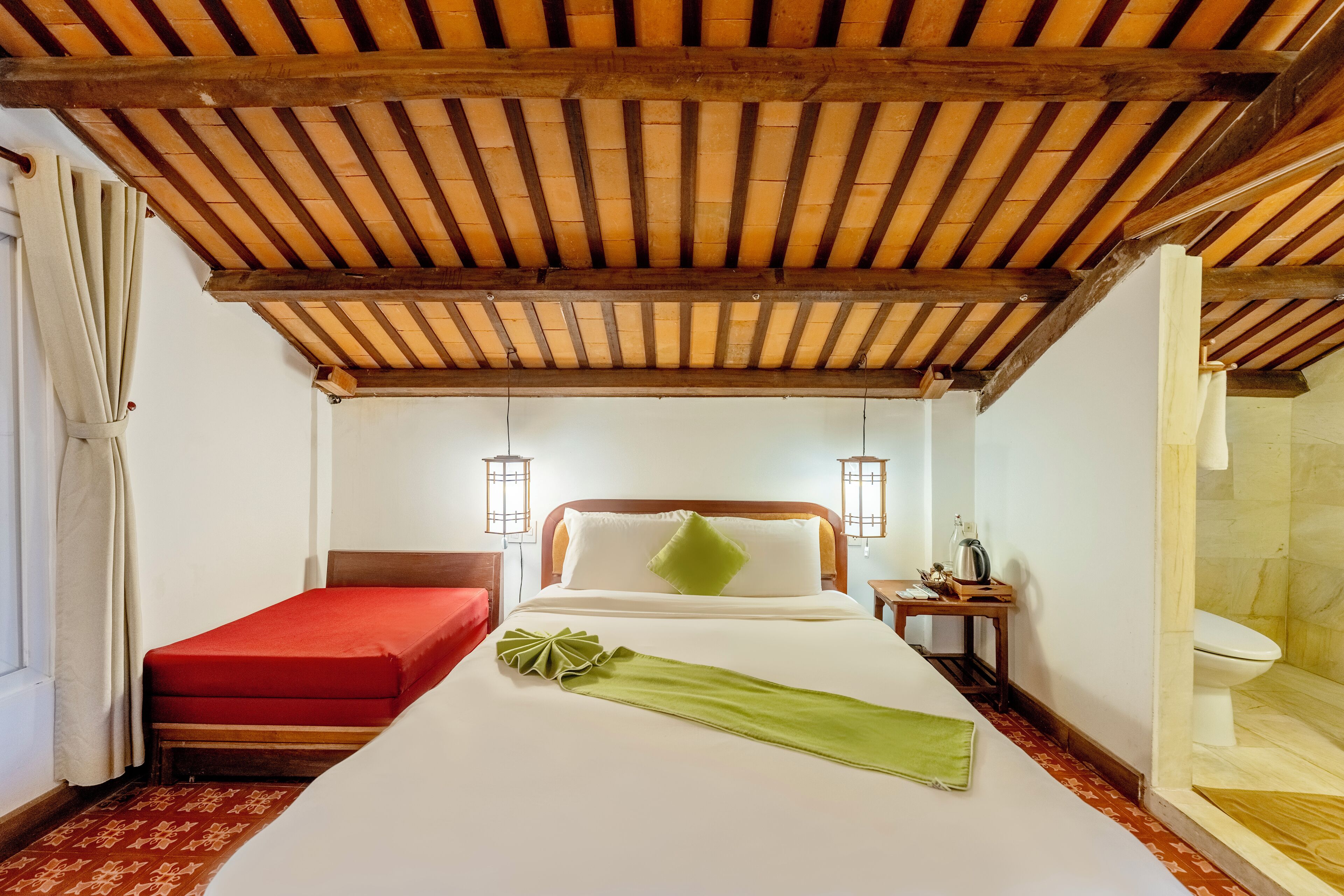 A hotel room with a wooden ceiling, a bed with a red mattress, a green throw, two bedside tables, and a small table with a kettle.