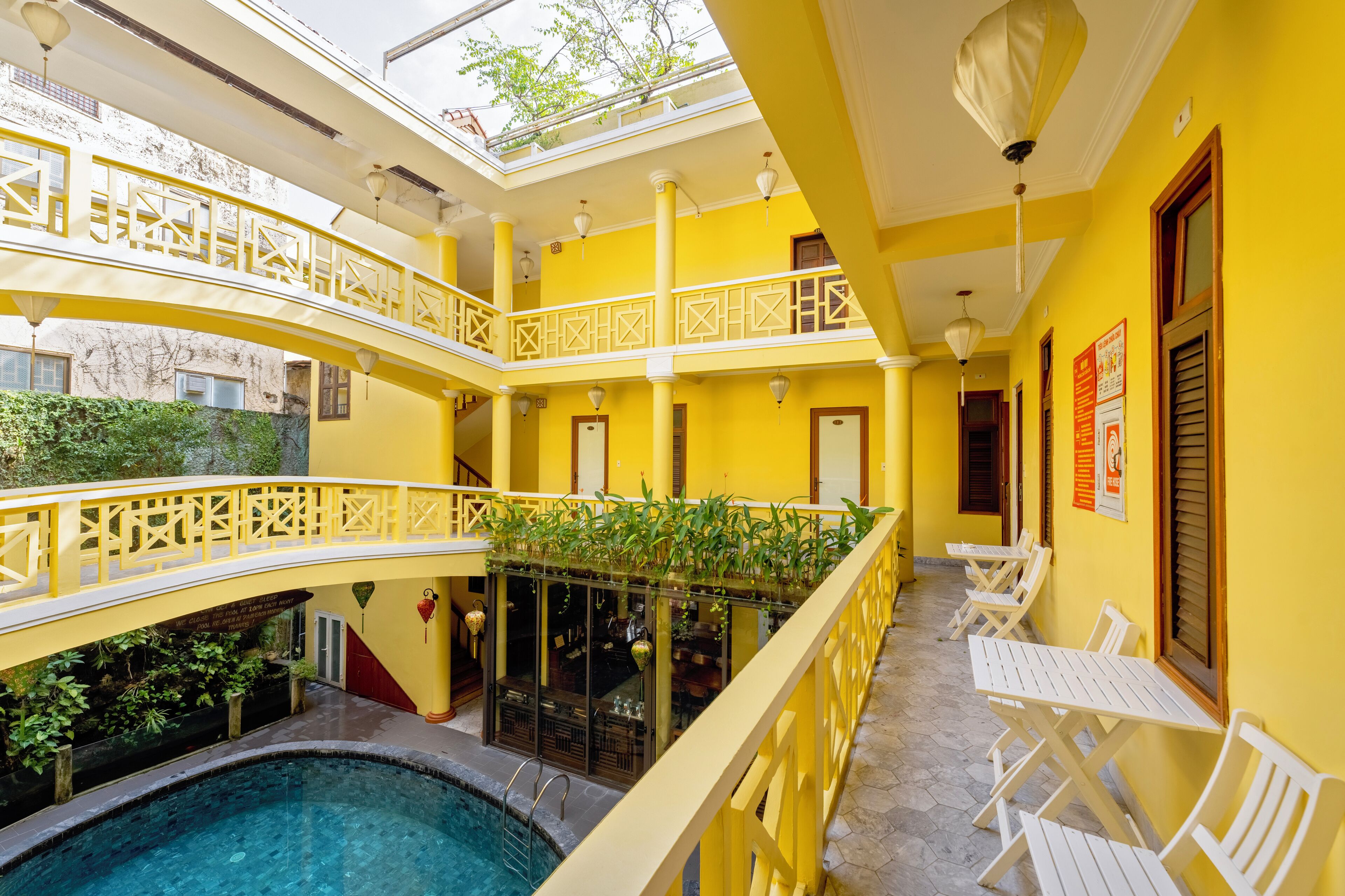 A multi-level yellow building with a pool, outdoor seating, and a covered area with plants.