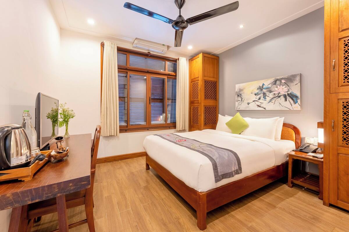 A hotel room with a wooden bed, a desk with a kettle, a plant, and a ceiling fan.