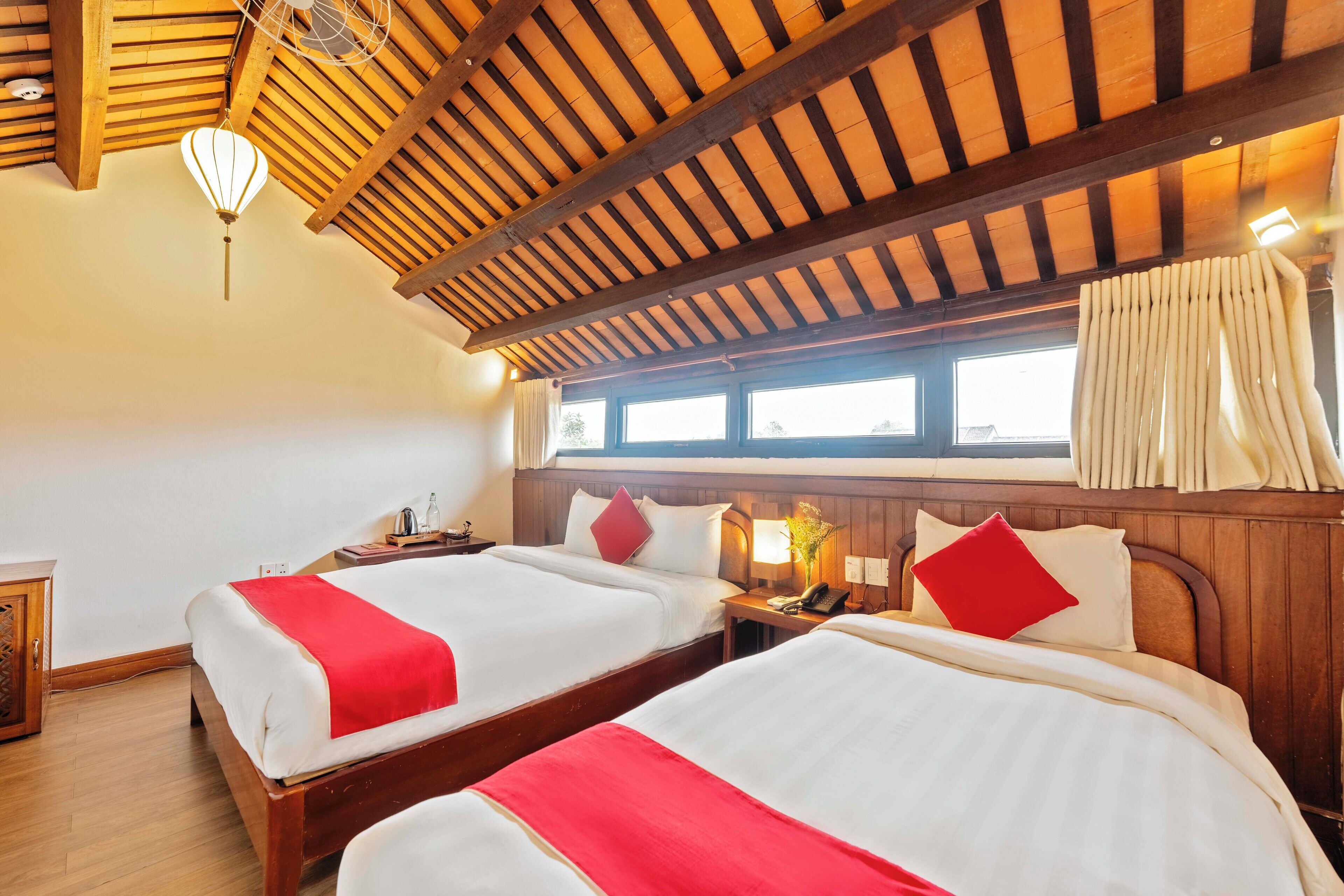 A hotel room with two beds, wooden beams on the ceiling, a window with curtains, and a bedside table with a lamp.