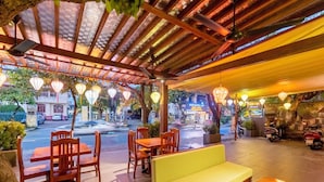 Un espacio al aire libre con mesas y sillas de madera, un banco verde con cojines y faroles colgantes.