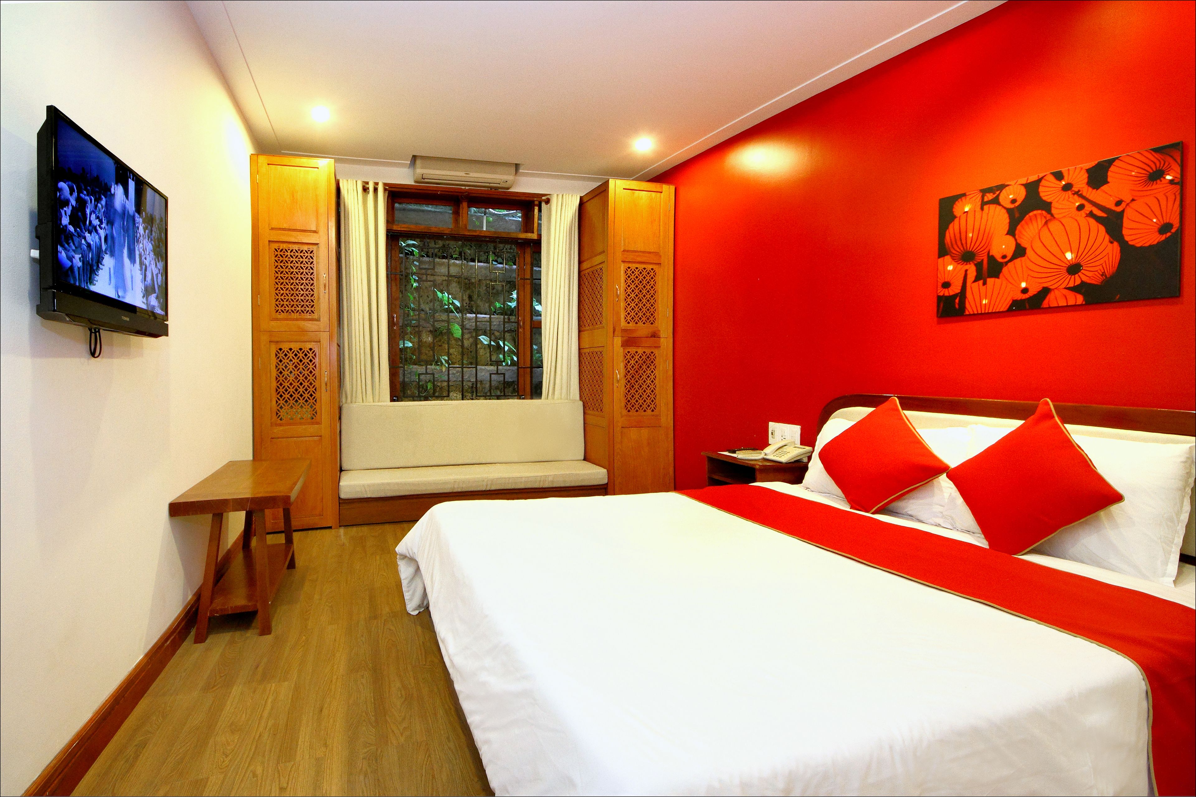 A hotel room with a bed, a television, a window with curtains, and a red wall with a floral painting.